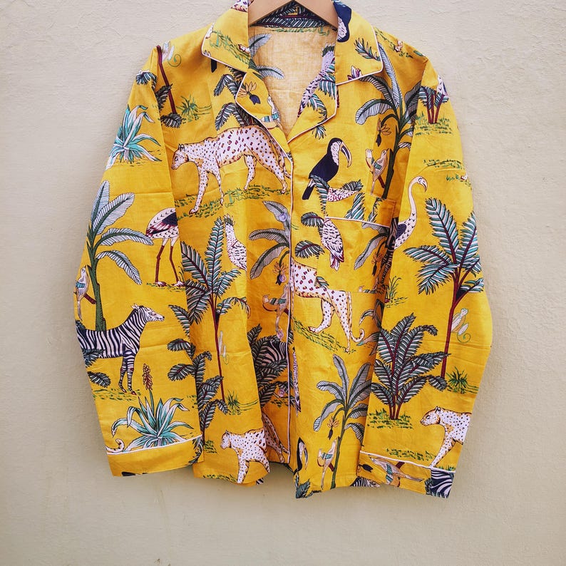 Pu&ograve; includere: Camicia da pigiama gialla con bottoni e stampa a tema giungla. Il design presenta zebre, leopardi, uccelli e palme. La camicia ha finiture bianche ed &egrave; appesa a un appendiabiti di legno.