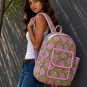 May include: A green and pink patterned backpack with a front pocket and pink trim. The backpack has padded shoulder straps and a top handle. The bag is made of a quilted fabric. The backpack is being worn over the shoulder.