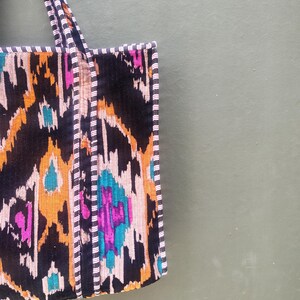 May include: A tote bag featuring a vibrant ikat pattern in black, orange, magenta, and teal. The bag has two matching fabric handles and is finished with a black and white pinstriped trim along the edges and the straps for a structured look.