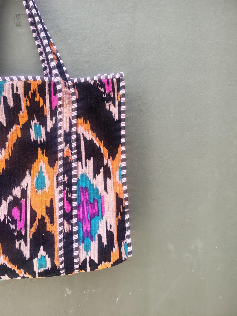 May include: A tote bag featuring a vibrant ikat pattern in black, orange, magenta, and teal. The bag has two matching fabric handles and is finished with a black and white pinstriped trim along the edges and the straps for a structured look.