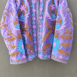 May include: A purple velvet jacket with intricate embroidery in shades of brown, teal, and pink. The jacket features a high collar and a full zipper. The lining is striped. This jacket is a unique piece of outerwear.