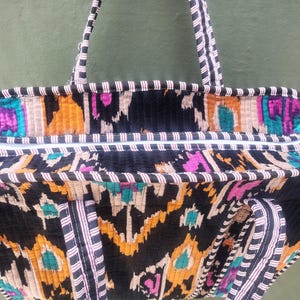 May include: A quilted tote bag featuring a vibrant ikat pattern in black, orange, teal, and magenta. The bag includes two striped fabric handles and a top zipper closure with a white and black striped trim along the upper edge and handles.