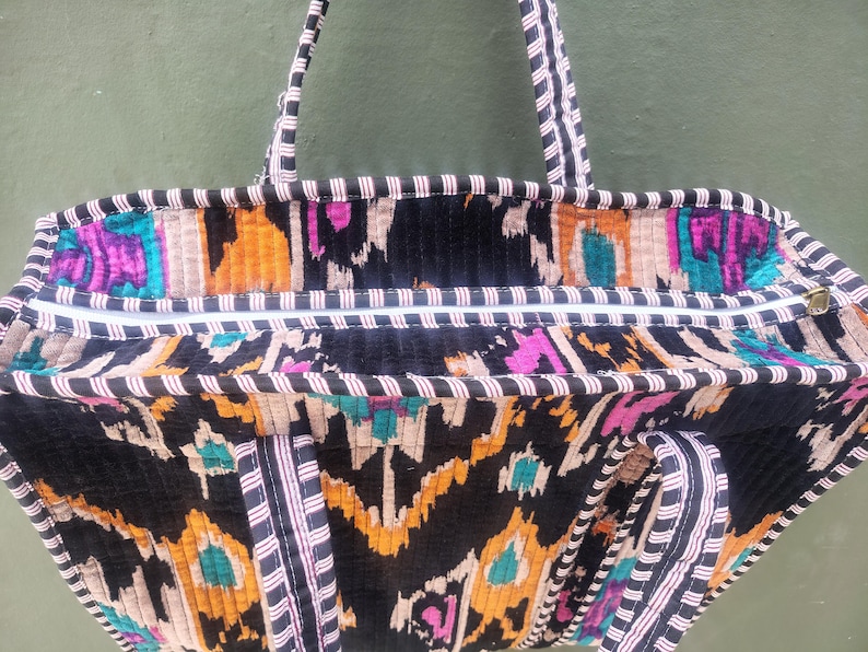 May include: A quilted tote bag featuring a vibrant ikat pattern in black, orange, teal, and magenta. The bag includes two striped fabric handles and a top zipper closure with a white and black striped trim along the upper edge and handles.