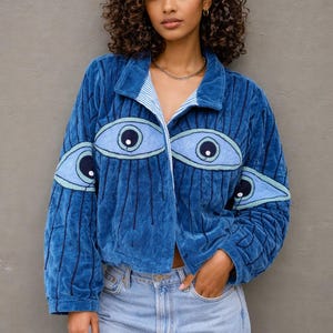 Op de afbeelding: Blauw fluwelen jasje met oogvormige applicaties. De jas heeft een kraag en verticale geribbelde details. De voering van de kraag is gestreept. Het model draagt een lichtblauwe jeans.