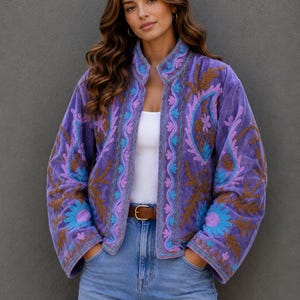 May include: A purple velvet jacket with intricate embroidery in shades of blue, brown, and purple. The jacket has a stand-up collar and is worn with a brown belt and blue jeans. The jacket's design features floral and paisley patterns.