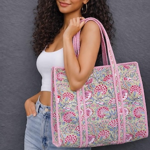 May include: A floral tote bag in pink, red, and blue on a light green background. The bag has pink trim and handles. The bag is held by a person wearing a white top and blue jeans.