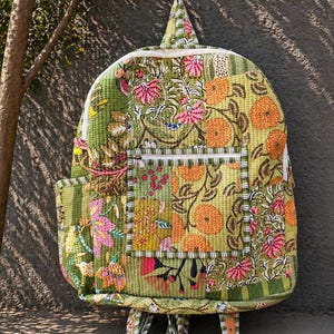 Könnte beinhalten: Ein gesteppter Rucksack mit einem lebendigen Blumenmuster in Grün-, Rosa- und Orangetönen. Der Rucksack hat eine Vordertasche, einen oberen Griff und verstellbare Träger. Das Design umfasst eine Mischung aus floralen und geometrischen Mustern.