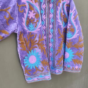 May include: A purple velvet jacket with intricate embroidery in shades of brown, blue, and purple. The jacket features a high collar and a striped lining. Floral and paisley patterns adorn the front and sleeves.