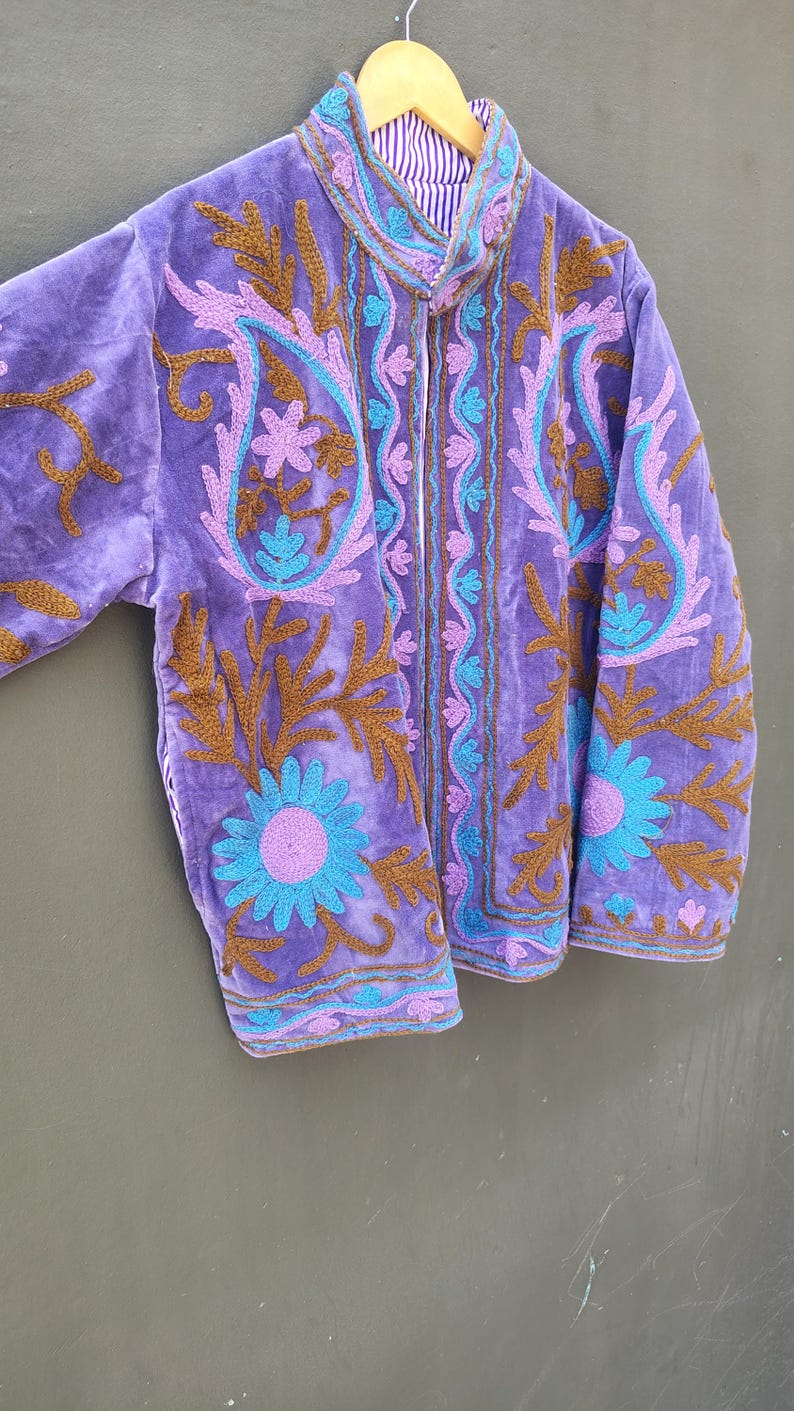 May include: A purple velvet jacket with intricate embroidery in shades of brown, blue, and purple. The jacket features a high collar and a striped lining. Floral and paisley patterns adorn the front and sleeves.