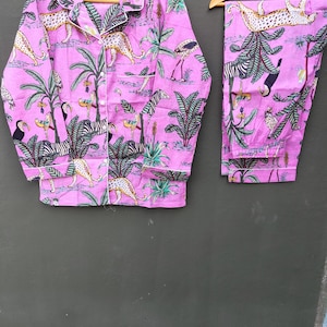 Puede incluir: Un conjunto de pijama rosa con estampado de animales de la jungla, incluyendo cebras, leopardos y tucanes. La camisa de manga larga y el pantal&oacute;n se muestran en perchas de madera. El conjunto tiene ribetes blancos.