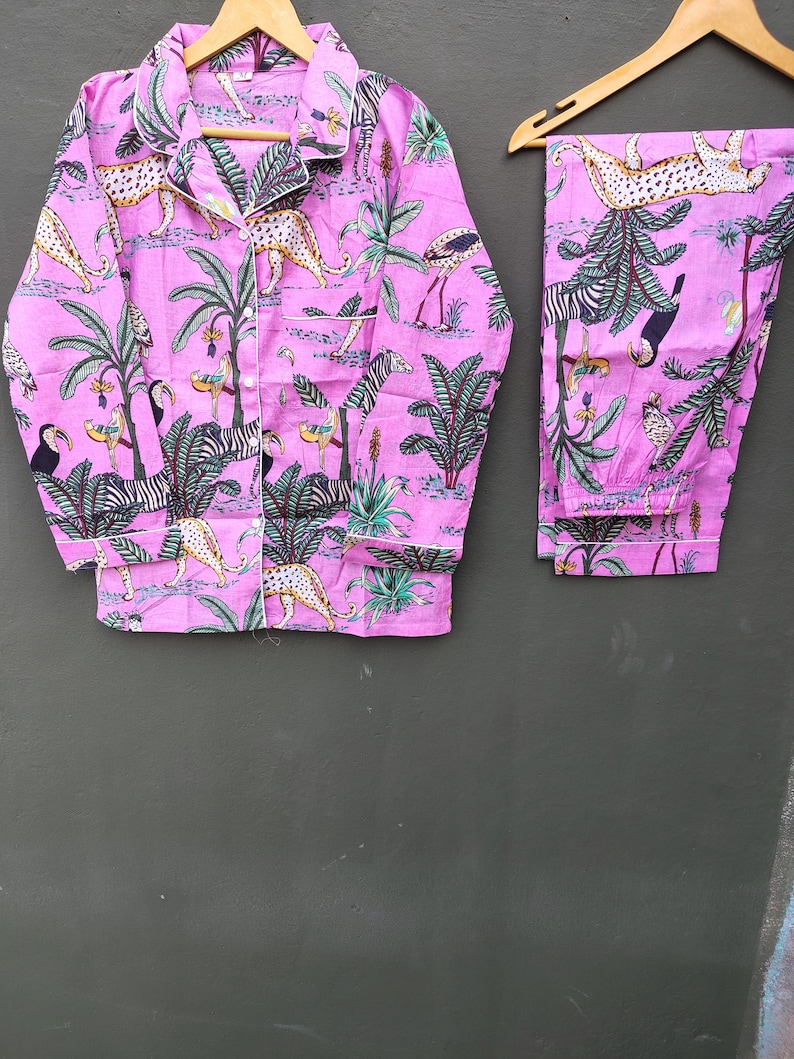 Puede incluir: Un conjunto de pijama rosa con estampado de animales de la jungla, incluyendo cebras, leopardos y tucanes. La camisa de manga larga y el pantal&oacute;n se muestran en perchas de madera. El conjunto tiene ribetes blancos.