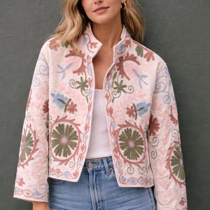 Peut inclure: Veste crème brodée avec un col montant. La veste présente des motifs floraux et de feuilles dans des tons de rose, vert et bleu. La veste est portée ouverte, révélant un haut blanc et un jean bleu.