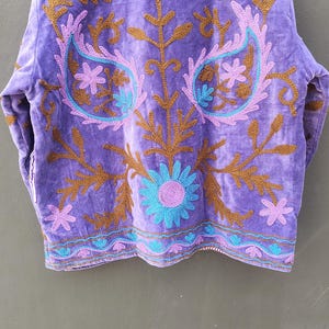 May include: A purple velvet jacket with intricate embroidery. The jacket features brown, teal, and lavender floral and paisley designs. The jacket is hanging on a wooden hanger against a neutral background.
