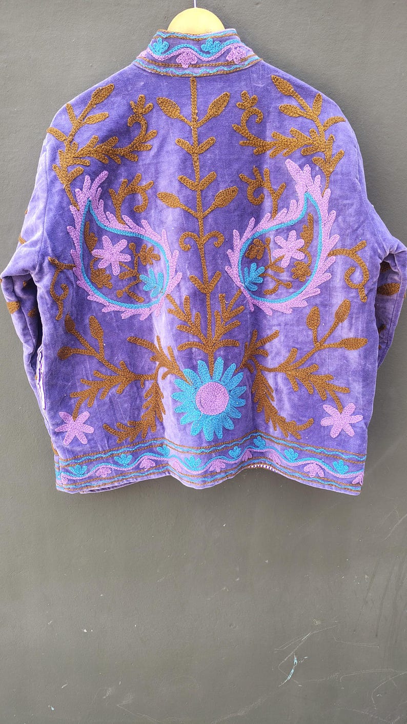 May include: A purple velvet jacket with intricate embroidery. The jacket features brown, teal, and lavender floral and paisley designs. The jacket is hanging on a wooden hanger against a neutral background.