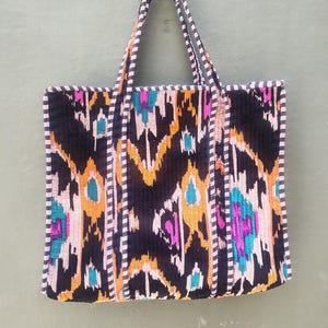 May include: A rectangular tote bag featuring a black, orange, teal, and magenta ikat pattern. The bag has two long shoulder straps and edges trimmed with a black and white striped fabric. The overall design is symmetrical with a quilted texture.