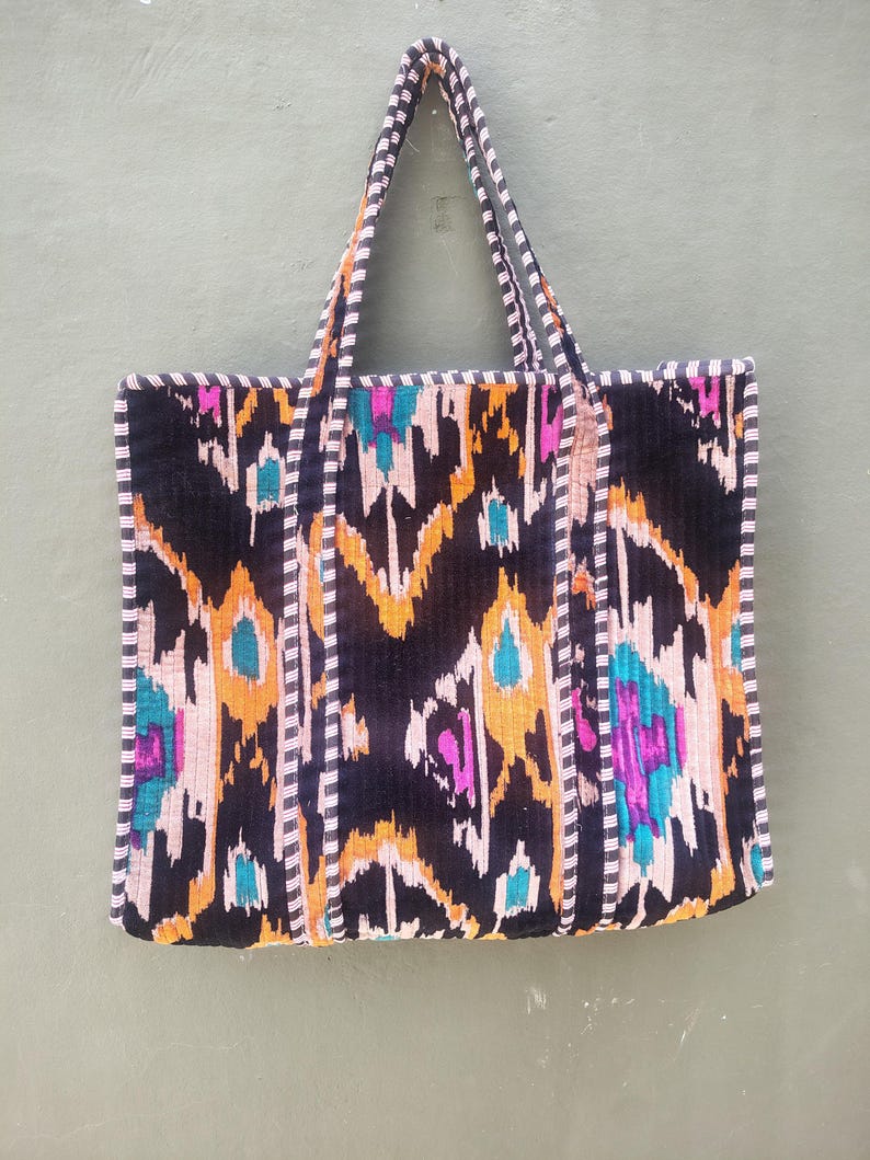 May include: A rectangular tote bag featuring a black, orange, teal, and magenta ikat pattern. The bag has two long shoulder straps and edges trimmed with a black and white striped fabric. The overall design is symmetrical with a quilted texture.
