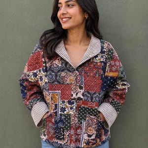 Op de afbeelding: Een gewatteerd jack met een patchworkontwerp met diverse bloemmotieven en geometrische patronen in tinten rood, blauw, groen en geel. Het jack heeft een omgeslagen kraag en manchetten met een wit en bruin krijtstreepmotief en twee zakken.