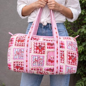 May include: A pink and white quilted duffel bag with a repeating pattern of cherries and bows. The bag has pink handles and trim, and a rectangular shape. The bag is held by a person wearing a white shirt and blue jeans.