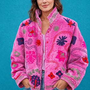 Pode incluir: Casaco rosa vibrante com bordados florais coloridos. O casaco apresenta uma gola alta e é adornado com vários desenhos florais bordados em tons de vermelho, roxo e verde. Usado sobre uma camisa listrada e jeans azul.