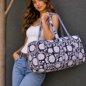 Könnte beinhalten: Eine marineblaue und weiße Reisetasche mit floralem Muster und gestreiften Griffen. Die Tasche ist gesteppt und zylindrisch. Die Person trägt ein weißes Tanktop und blaue Jeans. Das Design der Tasche zeigt ein Blumenmuster.