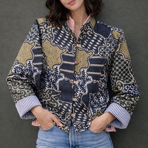 Könnte beinhalten: Gemusterte Jacke mit Stehkragen und langen Ärmeln. Die Jacke hat eine Mischung aus marineblauen, olivgrünen und weißen Mustern. Die Manschetten sind mit blau-weißen Streifen gefüttert. Die Jacke wird mit hellblauen Jeans kombiniert.