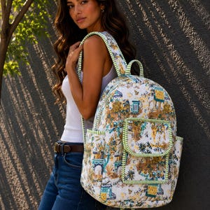 May include: A white backpack with a colorful floral and architectural pattern. The backpack has green trim and straps, and two front pockets. The straps are striped with green and white. The backpack is being worn over the shoulder.