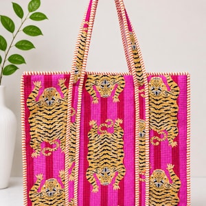 Könnte beinhalten: Eine gesteppte Tragetasche mit einem leuchtend pinkfarbenen Hintergrund mit vertikalen Streifen und einem sich wiederholenden Muster aus gelben und schwarzen Tigern. Die Tasche hat zwei lange Henkel mit orange-weiß gestreiftem Besatz.