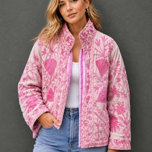 Pode incluir: Um casaco acolchoado rosa claro com bordados florais em magenta vibrante por todo o tecido. O casaco tem gola alta, mangas compridas e bainha reta, usado sobre uma blusa branca simples e calças de ganga azul claro.