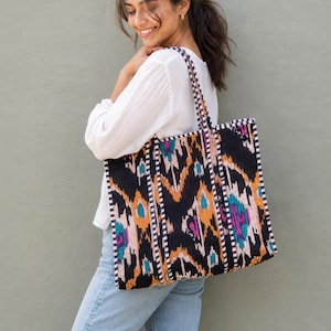 May include: A large rectangular tote bag featuring a black base with a vibrant ikat pattern in shades of orange, teal, and magenta. The bag includes two shoulder straps with a black and white checkered trim that matches the bag's side edges.