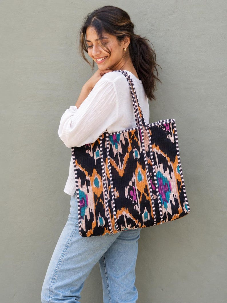 May include: A large rectangular tote bag featuring a black base with a vibrant ikat pattern in shades of orange, teal, and magenta. The bag includes two shoulder straps with a black and white checkered trim that matches the bag's side edges.