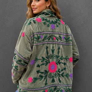 Pode incluir: Uma jaqueta de veludo verde-oliva com bordados intrincados em verde escuro, rosa brilhante e roxo. O design inclui motivos florais e faixas horizontais de costura nas costas e mangas, com gola alta e mangas compridas.