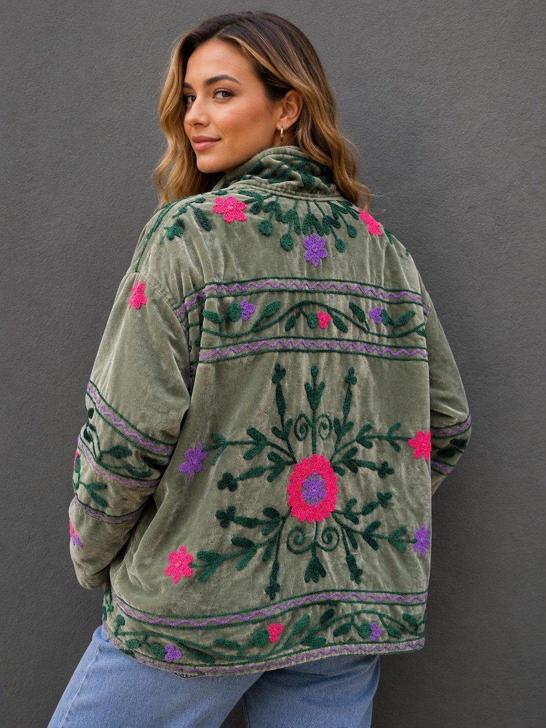 Pode incluir: Uma jaqueta de veludo verde-oliva com bordados intrincados em verde escuro, rosa brilhante e roxo. O design inclui motivos florais e faixas horizontais de costura nas costas e mangas, com gola alta e mangas compridas.