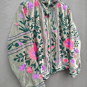 Pode incluir: Uma jaqueta de veludo verde s&aacute;lvia com bordados intrincados em padr&otilde;es florais verde-escuro, rosa choque e roxo. A jaqueta tem gola alta, mangas compridas e abertura frontal, com um forro listrado vis&iacute;vel na &aacute;rea do pesco&ccedil;o.