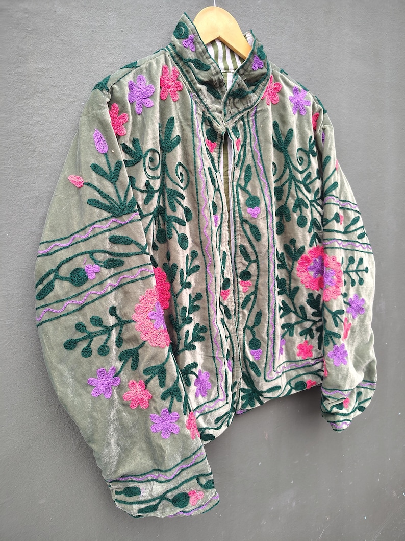 Pode incluir: Uma jaqueta de veludo verde s&aacute;lvia com bordados intrincados em padr&otilde;es florais verde-escuro, rosa choque e roxo. A jaqueta tem gola alta, mangas compridas e abertura frontal, com um forro listrado vis&iacute;vel na &aacute;rea do pesco&ccedil;o.