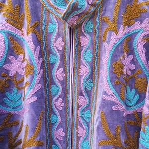 May include: A lavender velvet jacket with intricate embroidery in teal, gold, and purple. The jacket features a high collar and a striped lining. The floral and vine-like patterns create a bohemian aesthetic. The jacket is hanging on a wooden hanger.