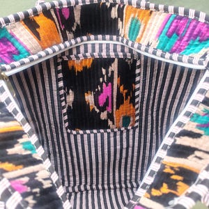 May include: A tote bag featuring a black and white striped interior lining and a zippered pocket. The upper rim and the pocket are decorated with a vibrant ikat pattern in shades of magenta, orange, teal, and black. It includes two fabric handles.