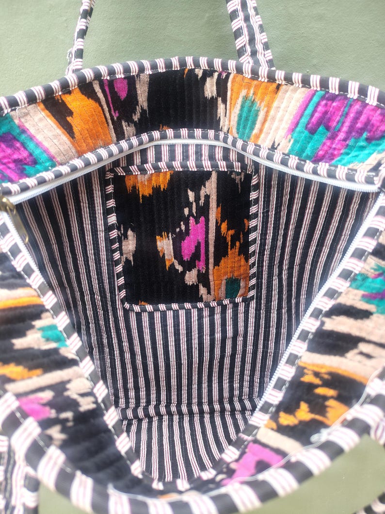 May include: A tote bag featuring a black and white striped interior lining and a zippered pocket. The upper rim and the pocket are decorated with a vibrant ikat pattern in shades of magenta, orange, teal, and black. It includes two fabric handles.