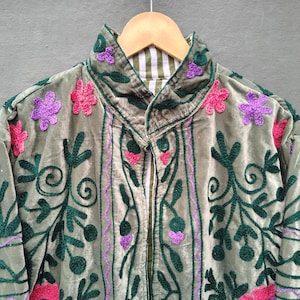 Pode incluir: Uma jaqueta de veludo verde s&aacute;lvia com padr&otilde;es intrincados de vinhas bordadas em verde escuro e detalhes florais vibrantes em rosa e roxo. A pe&ccedil;a possui uma gola alta e um forro listrado de branco e verde vis&iacute;vel na abertura do pesco&ccedil;o.