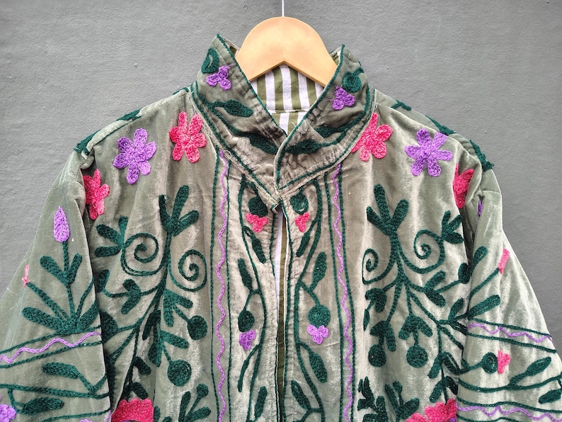 Pode incluir: Uma jaqueta de veludo verde s&aacute;lvia com padr&otilde;es intrincados de vinhas bordadas em verde escuro e detalhes florais vibrantes em rosa e roxo. A pe&ccedil;a possui uma gola alta e um forro listrado de branco e verde vis&iacute;vel na abertura do pesco&ccedil;o.