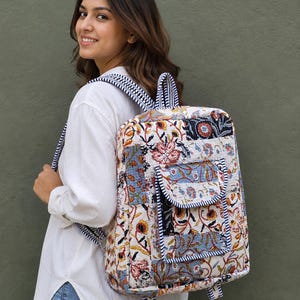 Könnte beinhalten: Ein gesteppter Stoffrucksack mit einem mehrfarbigen Blumenmuster in Blau-, Orange- und Schwarztönen auf weißem Grund. Der Rucksack verfügt über eine Vordertasche mit Klappe, einen Tragegriff oben und verstellbare Schultergurte mit blau-weißen Streifen.