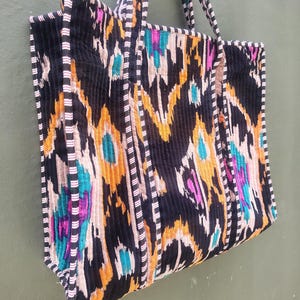 May include: A quilted tote bag featuring a black, orange, and teal ikat pattern. The bag has two shoulder straps and black and white striped piping along the edges. The fabric has a vertical ribbed texture throughout the exterior surface.