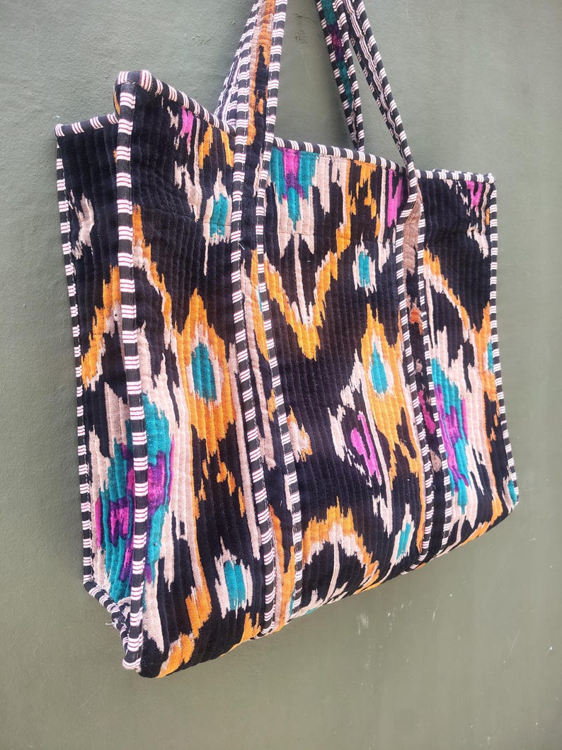 May include: A quilted tote bag featuring a black, orange, and teal ikat pattern. The bag has two shoulder straps and black and white striped piping along the edges. The fabric has a vertical ribbed texture throughout the exterior surface.