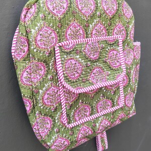 May include: A quilted backpack in olive green with a repeating pink floral pattern. The backpack has a front pocket and is trimmed with pink and white striped fabric. The straps and handle are also striped.
