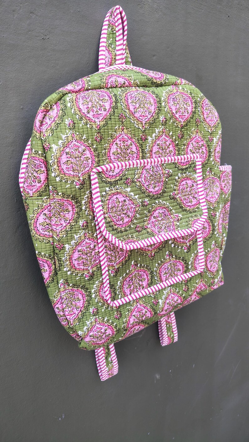 May include: A quilted backpack in olive green with a repeating pink floral pattern. The backpack has a front pocket and is trimmed with pink and white striped fabric. The straps and handle are also striped.