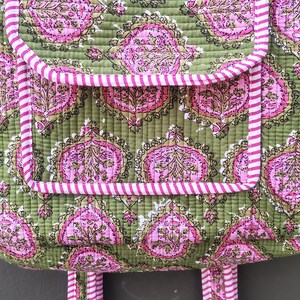 May include: Close-up of a quilted tote bag with a green background and a pink floral pattern. The bag has two pockets and a pink and white striped trim. The handles are also pink and white striped.