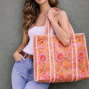May include: A large tote bag with a floral pattern in shades of pink, orange, and yellow. The bag has a rectangular shape with sturdy handles and a matching trim. The background is a solid grey-green.