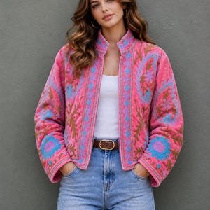 Può includere: Giacca trapuntata rosa con ricami floreali blu, viola e oro. La giacca ha un colletto alla coreana e viene indossata con un top bianco e jeans blu. Una cintura marrone segna il punto vita.