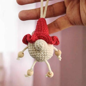 May include: A handmade crochet gnome ornament with a red hat and beige body. The gnome has wooden bead accents for arms and legs, and is suspended by a cream-colored string. The text "Woolly Smile" is in the upper left corner.