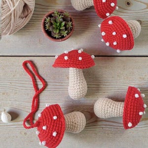 Op de afbeelding: Een verzameling handgemaakte gehaakte paddenstoelen met rode hoeden en witte stippen, en beige stelen. De paddenstoelen zijn gerangschikt op een licht houten oppervlak, samen met een kleine vetplant in pot en een bol garen. De paddenstoelen variëren in grootte en vorm.