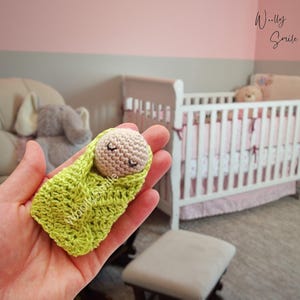 May include: A small crocheted baby doll, wrapped in a green blanket, with closed eyes and a small smile. The doll is being held in a person's hand. The background shows a crib with pink and white bedding.